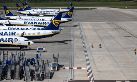 Three people walk on the runway amongst parked and temporarily out of service Ryanair passenger aircraft at Stansted Airport.