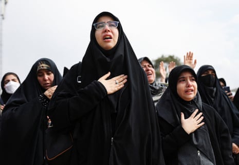 Crying women dressed in black and wearing dark hijabs stand with one hand on their chest.