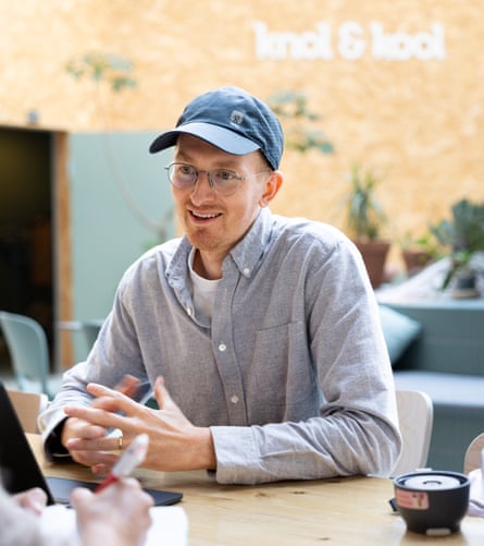 A man wearing a blue baseball cap and a grey shirt sitting at a desk with a brick wall with the words ‘knol & kool’ behind him