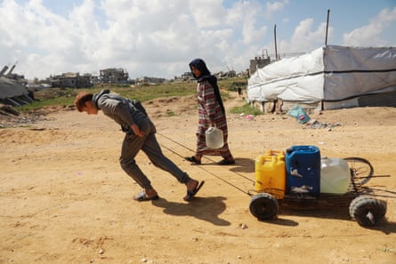 A young man pulls a platform trolley loaded with plastic jerry cans along a dirt road as a woman carrying containers walks next to him