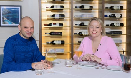 A bald man wearing a bright blue patterned jumper, and a blond woman wearing a pink cardigan, sitting at a restaurant table smiling
