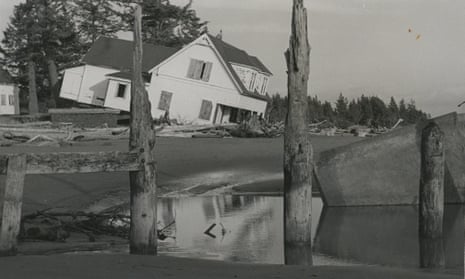 A black and white image of a beachfront house that has come loose from its foundation and tipped over onto the sandy shore.