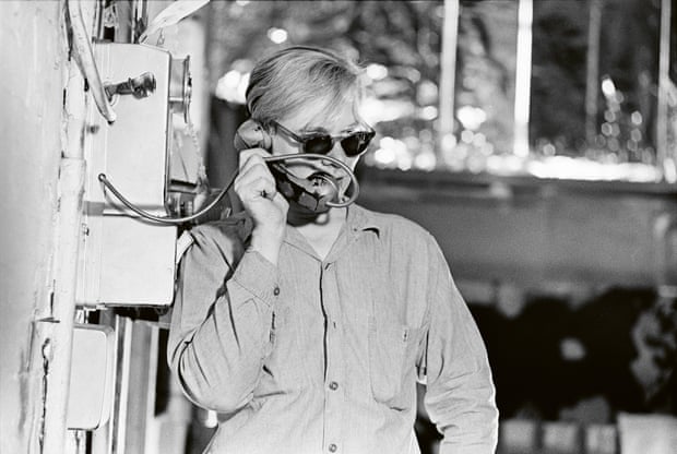 Andy Warhol wearing a shirt and sunglasses, speaking on a phone on the wall of an industrial building