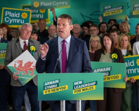 Rhun ap Iorwerth gestures while speaking at a podium with Plaid Cymru campaign signs behind him.
