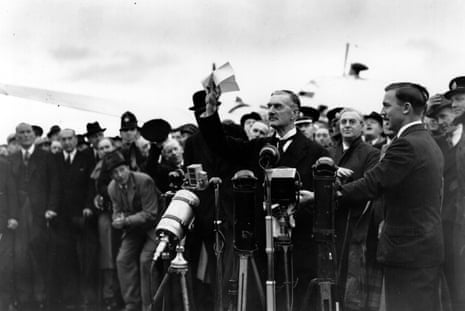 Neville Chamberlain standing at a microphone holding a piece of paper in the air among a crowd at an airport