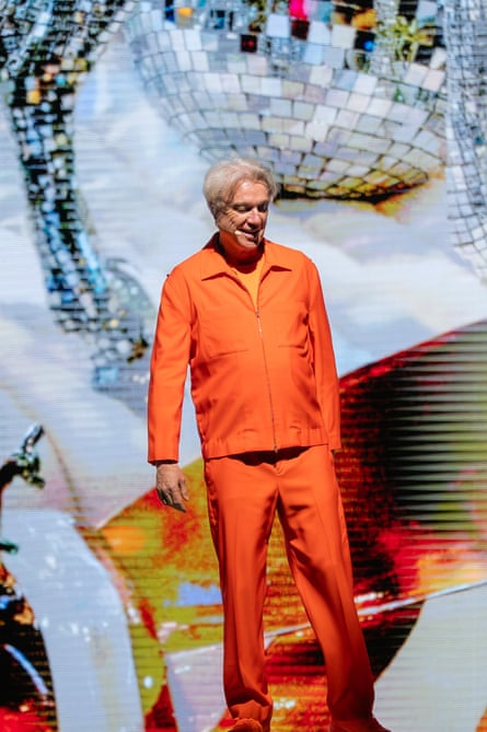 David Byrne wears an orange jacket and pants and stands in front of a mirrorball