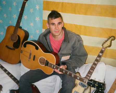 Rostam Batmanglij holds an acoustic guitar while sitting between two other guitars against a backdrop of a faded American flag
