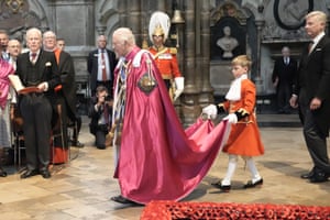 King Charles walks in Westminster Abbey wearing a dark pink robe while a boy in a red frock coat carries the robe’s train