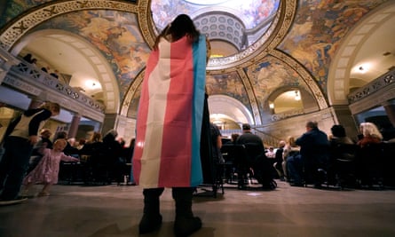 A counter-protester stands at back of church with painted ceiling and wears a transgender flag as people sit in church, where they are supporting a ban on gender-affirming healthcare legislation