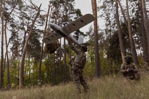 A soldier standing a field in front of woodland points what looks like a small aircraft skywards while another behind him operates a remote control