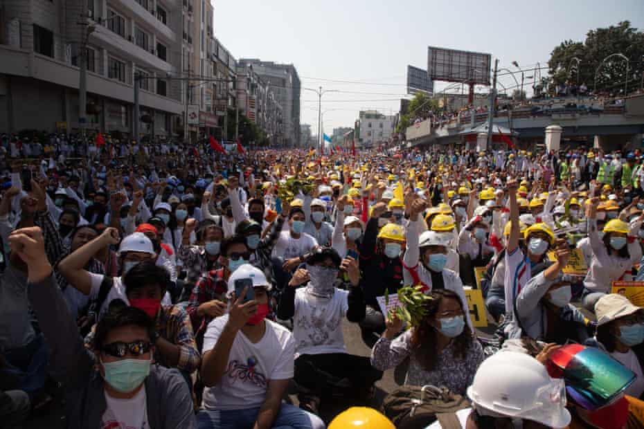 Large numbers of seated protesters with raised fists in front of Mandalay Central station