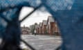 Glimpse through a torn fabric covering of a fence into an empty square lined with old red brick barracks accommodation, with only one person visible in the aquare