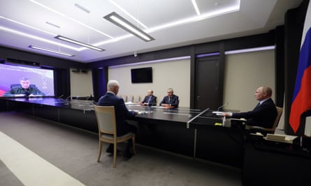 A modern conference room with Putin and three other men sitting at a long table, looking at a large screen at one end of the room with an image of Gerasimov