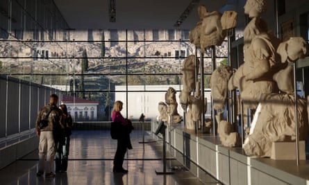 People look at sculptures on display in the museum, with the Acropolis visible through windows behind