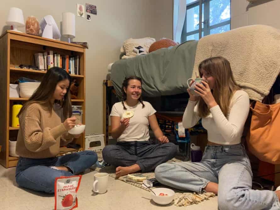 Three female Pomona college students sit on the floor of their dorm room eating breakfast. The student at the far left eats from a bowl, the center student smiles as she holds a bagel, and the student at right sips from a mug. behind them is a lofted bed and a bookcase.