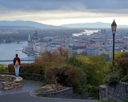 A man with a backpack looks out over a city with a winding river on a cloudy, overcast day