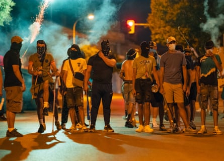 Men wearing masks and balaclavas hold chains, sticks and baseball bats; they are dressed in shorts and T-shirts and stand in a road under street lights at night; fireworks are exploding in the background.