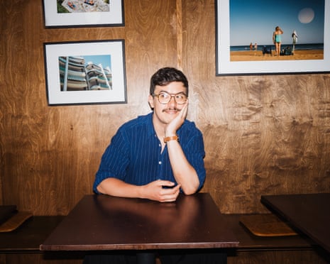 Comedian Phil Wang sits at a wooden table, resting his chin on his hand, with framed photographs on a wood-panelled wall behind him
