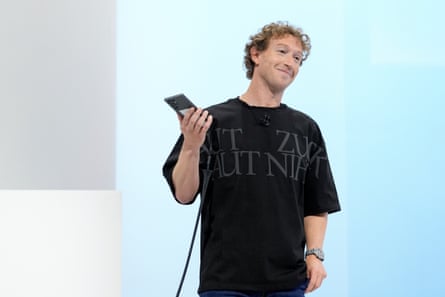 Марк Цукерберг у чорній футболці з малюнком усміхається на сцені, тримаючи в руках пристрій