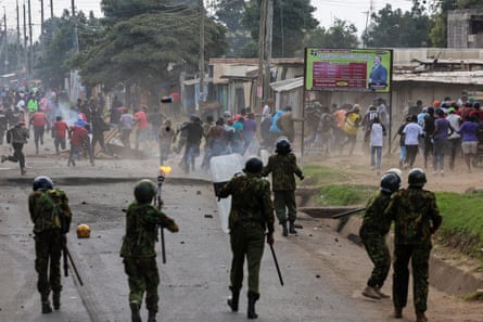 Police, some with riot shields, fire at a crowd of protesters who are running away.