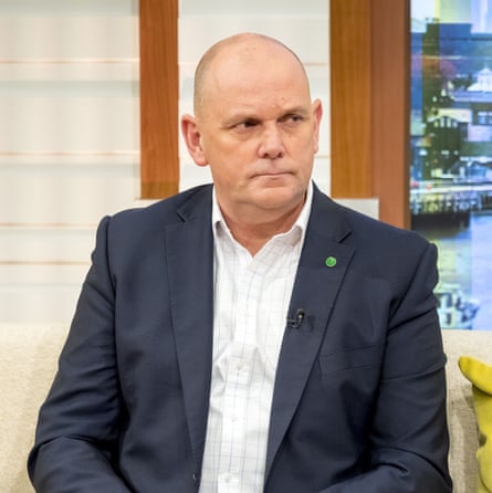 he sits in a TV studio wearing an open-necked white shirt and dark blue jacket; he is middle-aged, bald, and looks stern.