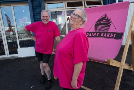 A man and a woman in bright-pink tops stand outside their cafe, smiling widely