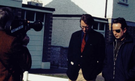 J Bowyer Bell, in a suit and tie and glasses, looks down as he walks next to Seán MacStíofáin, Provisional IRA chief of staff, who wears an eye patch – both walking past houses towards a cameraman with a camera on his shoulder