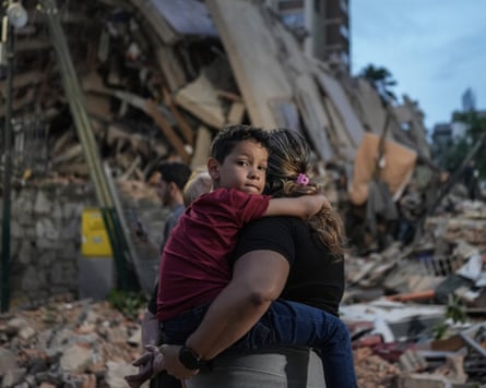 A child is carried in front of a destroyed building