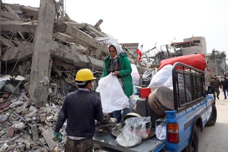 A woman in a green jacket and white hijab stands on the back of a blue pickup truck holding a white bin linger while a man with a yellow construction hat stands next to her. They are all next to the rubble and debris of ruined building.