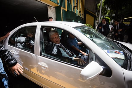 A grey-hared man in a suit driving a light-coloured car.