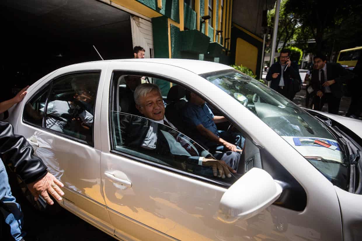 A grey-hared man in a suit driving a light-coloured car.