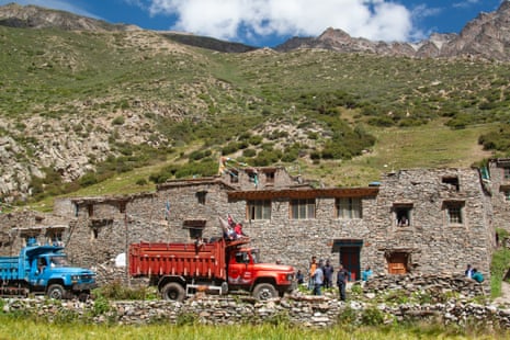 Villagers stand in a road next to a red and blue truck against the backdrop of a mountainous slope