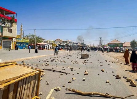 Debris is strewn across a road while smoke rises over buildings in the distance.