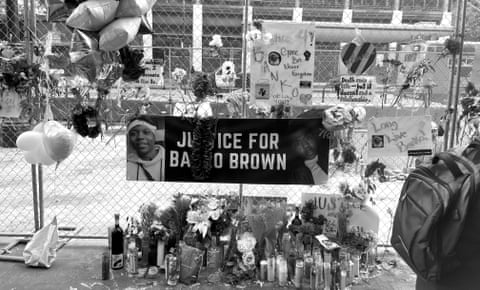 black and white photo shows pictures of brown on a sign that says 'justice for banko brown', on a chain link fence with flowers and messages