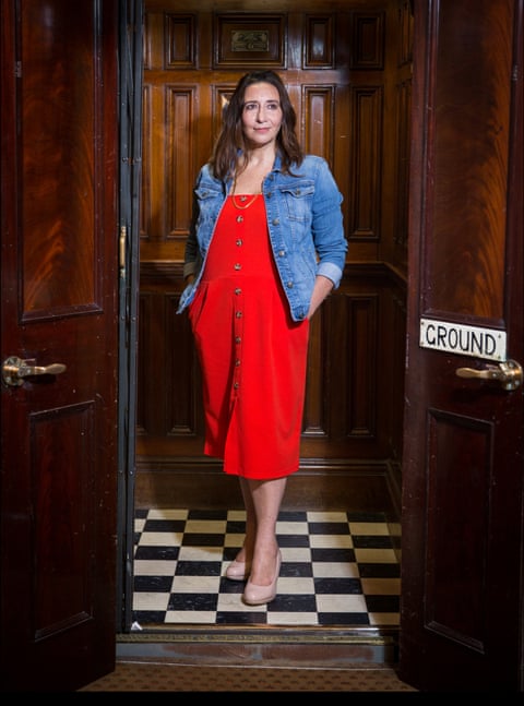 Bibi Lynch wearing a red dress and jean jacket with her hands in her pockets standing in a wood panelled lift