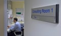 Doctor sitting at a desk, as viewed through an open door with a sign reading 'consulting room 1' on the wall outside