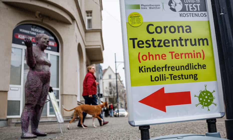 A woman in a red coat walking a dog walks past a street sign that says Corona Testzentrum