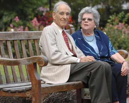 An older man in beige suit jacket and an older woman in a blue top sit on a bench.