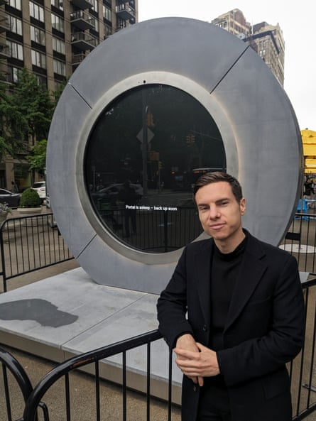 Man wearing black suit and black shirt leans against railing in front of grey circular structure