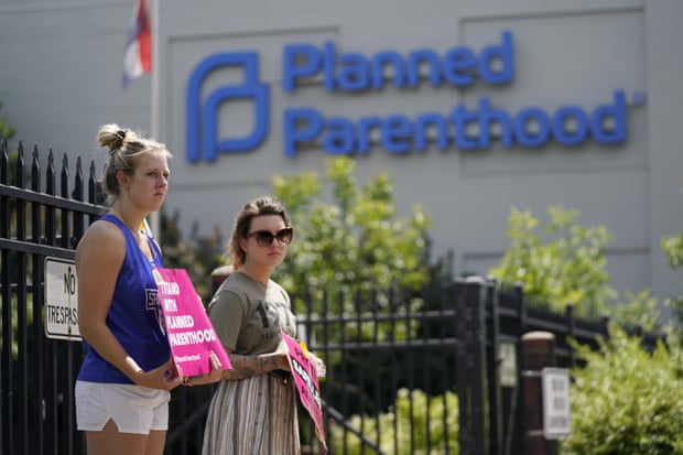 Two women holding pink signs in support of abortion rights stand outside the black iron fence of a Planned Parenthood building.