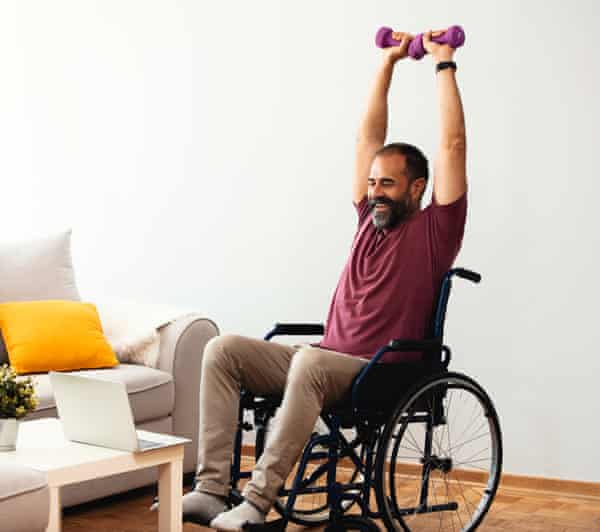 An elderly man in a wheelchair, smiling, lifts two dumbbells over his head