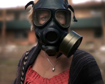 She looks at the camera, wearing a black gas mask