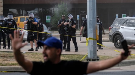 man in blue baseball cap stands with arms in the air as a group of officers in tactical gear behind him stand across street holding weapons