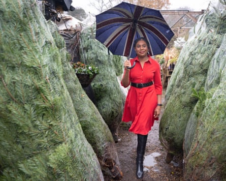 she is walking between a line of trees that are bagged up to be carried away. She wears a bright red dress, which contrasts with the green of the trees, and carries an open union jack umbrella above her head.