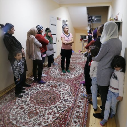 A group of women and children stand talking in a long room with a Persian carpet