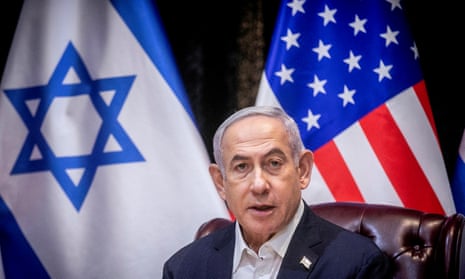 Benjamin Netanyahu pictured from the shoulders up, wearing a white shirt and dark jacket, with the Israeli and US flags visible behind him.