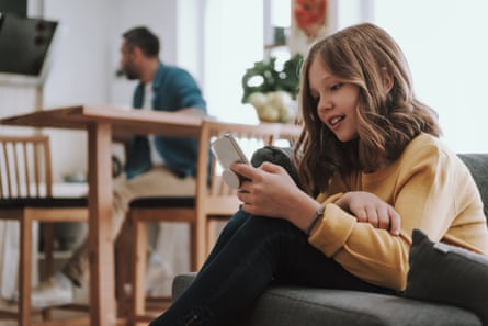 Girl with long fair hair looking at phone at home, sitting on a couch; her father is in the background at a table.