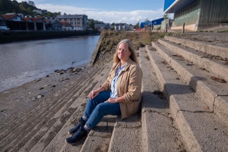 Hannah Pearson sits on stone steps by the riverside with the town in the background. She has long, straight fair hair and wears a sand-coloured cotton jacket and blue jeans.