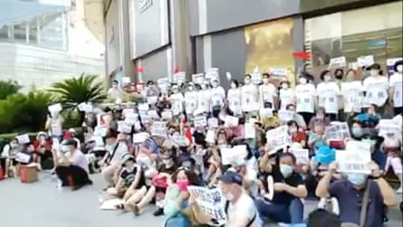 A large group of protesters sitting or standing outside an office building, many holding placards