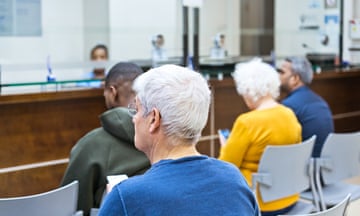 Posed by models Back view of multiracial group of people, many of them older, sitting in waiting room in hospital. Reception in the background.